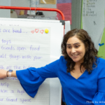A teacher sits next to a whiteboard in a classroom and points to a list of ways to be kind